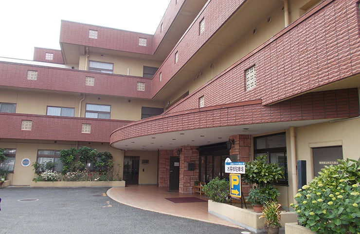 赤レンガ造りの介護施設の建物外観。曲線的な設計で、玄関アプローチと緑が配置されている。